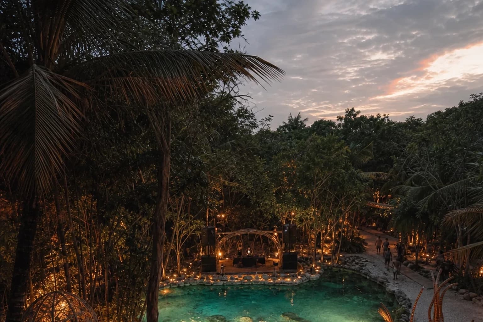 Open-air Zamna Tulum jungle venue at dusk with a cenote pool glowing at center stage, surrounded by tropical canopy and silho